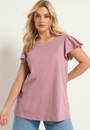 Blondynka w jasnofioletowej bluzce z krótkim rękawem i falbanami oraz jasnoniebieskich dżinsach, stojąca na jednolitym tle.