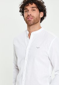 Mann mit lockigem Haar, der ein weißes, langärmeliges Hemd mit Knöpfen und einem kleinen gestickten Logo auf der Brust trägt, vor einem schlichten Hintergrund stehend.