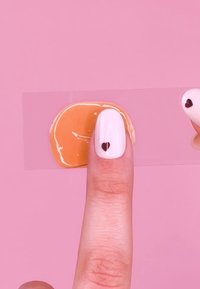 Nagellak in glanzend wit met een zwart hartontwerp, bedekt met een glanzende oranje gel op een vinger, tegen een roze achtergrond.