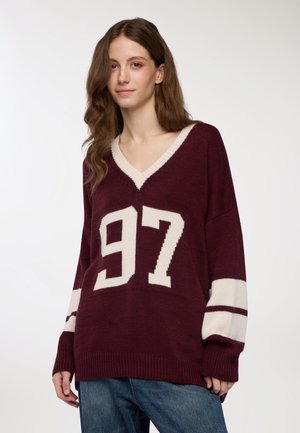 Giovane donna con lunghi capelli castani, che indossa un maglione a V colore bordeaux con la scritta bianca "97" e strisce sulle maniche, abbinato a jeans blu.