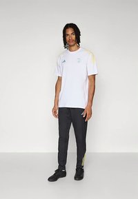 Bela športna majica s kratkimi rokavi z modrimi in rumenimi poudarki, kombinirana s črnimi zoženimi hlačami z rumenimi črtami in črnimi supergami.