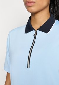 Svijetloplava polo majica s tamnoplavim ovratnikom, koja ima zatvaranje na patentni zatvarač sprijeda s dvije crne pruge i dekorativnim detaljem od hardvera.