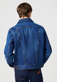 Modrá džínová bunda s klasickým střihem, knoflíkovými manžetami a dvojitými švy. Obsahuje zadní sedlo a kovové detaily.