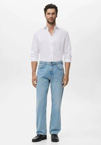 Man staat in een witte button-up shirt, lichtblauwe jeans en zwarte schoenen tegen een effen lichte achtergrond.