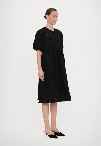 Black lace dress with a round neckline, short puff sleeves, and an A-line silhouette. Paired with black pointed flats.