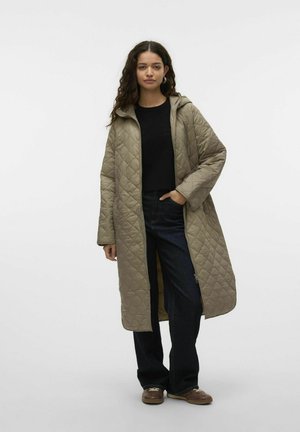 Manteau long matelassé en beige clair, doté d'une capuche, d'une fermeture éclair à l'avant et de fentes sur les côtés. Associé à un haut noir et un jean foncé.