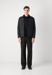 Schwarze gepolsterte Jacke mit Kragen, zwei Brusttaschen und Reißverschluss, kombiniert mit einem schwarzen Hemd mit Knöpfen und einer Hose.