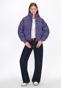 Fialová péřová bunda se vzorem zvířat přes celé tělo, vysokým límcem a elastickými manžetami, spárovaná s širokými tmavě modrými džínami a bílými botami.