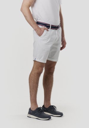 Bărbat purtând pantaloni scurți albi, un tricou alb, o curea bleumarin și pantofi casual bleumarin cu șireturi, stând cu o mână în buzunar.