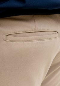 Gros plan sur un pantalon en tissu beige montrant une poche arrière horizontale avec des coutures détaillées et une ceinture noire au-dessus.