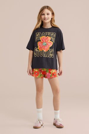 Mladé dievča stojí, oblečené v čiernom oversized tričku s veľkým oranžovým kvetom a nápisom „Flower Stories“, kvetinové šortky, biele ponožky a ružové tenisky.