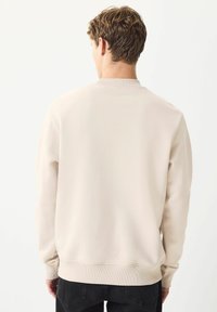 Man med kort lockigt hår som bär en enkel beige sweatshirt och svarta jeans, står vänd bort mot en vit bakgrund.