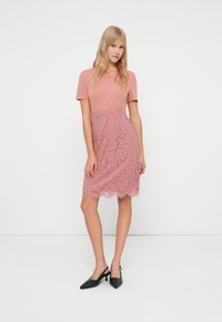 Vestido rosa com um corpete liso e uma saia de renda texturizada, com um bainha recortada e mangas curtas. Combinado com sapatos de salto pretos.