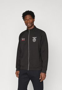 Juodas Zip-up džemperis su stovinčiu apykakle, kairėje pusėje puikuojasi emblema ir raudonas tekstas, pagamintas iš lygios, minkštos medžiagos.