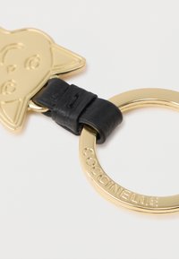 Kultainen metallinen avaimenperä, jossa on tyylitelty kissamalli; sisältää mustan nahkakiinnikkeen ja pyöreän avainrenkaan, jossa on kaiverrettuna "Coccinelle".