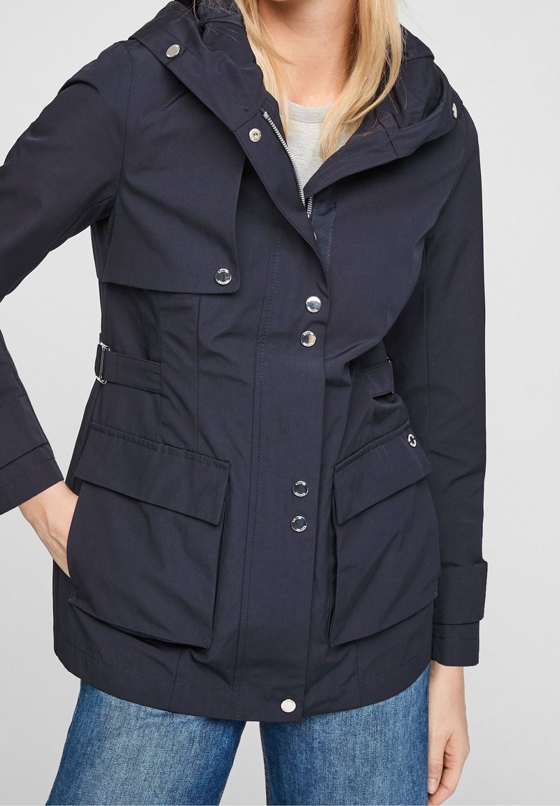 Veste imperméable bleu marine avec capuche, fermeture à boutons et à glissière, quatre poches avant, et ceinture de taille réglable pour une coupe ajustée.