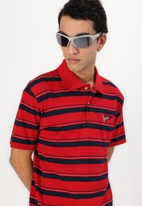 Crvena polo majica s tamnoplavim prugama, s kragnom i malim logotipom. Sive sunčane naočale upotpunjuju izgled. Glatka tekstura.