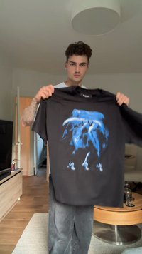 Nuori mies, jolla on tatuointeja, pitää mustaa T-paitaa, jossa on sininen abstrakti kuvio. Hän seisoo modernissa olohuoneessa, jossa on puulattia ja huonekalut.