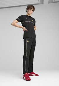 Sorte Puma t-shirt med hvide detaljer, sorte træningsbukser med gult logo og røde Puma sneakers. Modellen viser en atletisk pasform i en neutral baggrund.