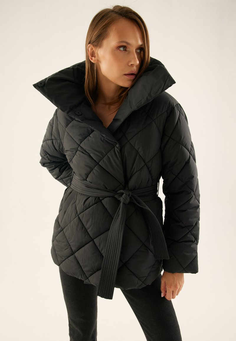 Kobieta ubrana w czarną pikowaną kurtkę z szerokim kołnierzem i paskiem związanym w talii, stojąca na jednolitym jasnym tle.