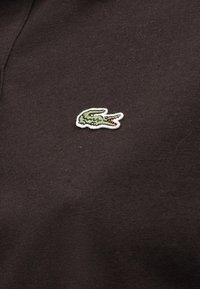 Logo de crocodile vert brodé avec une bouche rouge sur un tissu marron foncé, centré sur un vêtement.