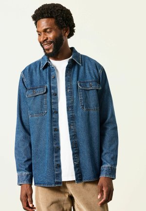 Mann lächelt und schaut nach unten, trägt eine blaue Jeansjacke über einem weißen Hemd und hellbraune Cordhosen vor schlichtem Hintergrund.