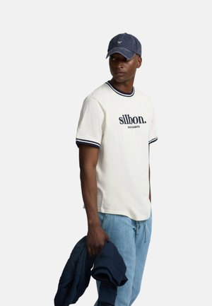Hombre con camiseta blanca Silbon con ribetes azul marino y gorra, sosteniendo una chaqueta azul marino, de pie sobre un fondo liso y claro.
