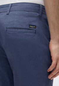 Παντελόνια σε ναυτικό μπλε χρώμα από λείο ύφασμα, με πίσω τσέπη και μια μικρή μαύρη ετικέτα με την επιγραφή "Scotch & Soda" στη μέση.