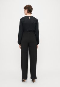 Blouse noire à manches longues avec un motif texturé et un détail en trou de serrure au dos, associée à un pantalon large noir et des chaussures à talons hauts noires.