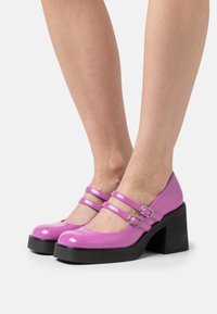 Chaussures à plateforme en cuir verni rose avec deux sangles à boucles réglables. Talon épais noir et design à bout rond. Texture lisse et brillante.