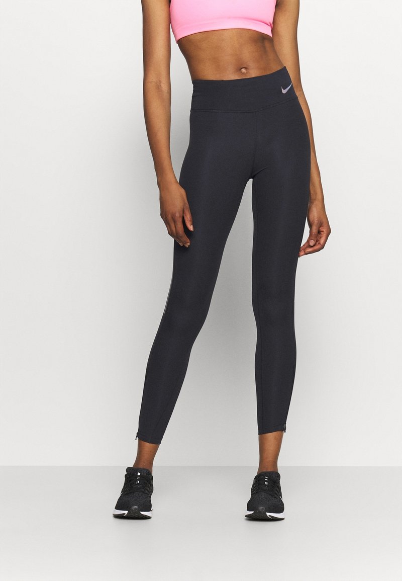 Leggings deportivos negros con una cintura alta, tobillos con cremallera y un pequeño logo morado. Llevados con un top corto rosa y zapatillas negras.