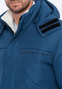 Modrá zateplená bunda s vysokým límcem, doplněná černými suchými zipy na ramenou a textem logo. Hladká, voděodolná tkanina.