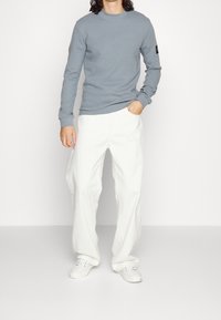 Graues Langarmshirt mit gerippter Textur, kombiniert mit weißen weit geschnittenen Hosen und weißen Sneakers. Schwarzes Logo-Patch am Ärmel.