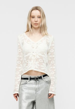 Mujer con una blusa blanca de crochet de manga larga con lazo delantero y pantalones plateados metálicos con cinturón negro, de pie frente a un fondo liso.