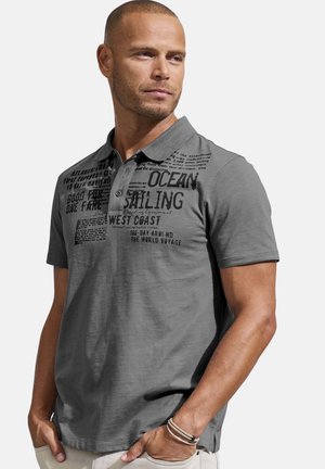 Mann in grauem Poloshirt mit schwarzem, segelthemenbezogenem Text steht mit den Händen in den Taschen und schaut seitlich vor schlichtem Hintergrund.