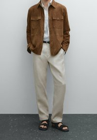 Veste-chemise en daim marron avec deux poches poitrine, chemise blanche à boutons, pantalon couleur crème et sandales noires à deux brides.