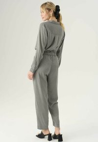 Grijze jumpsuit met een patroon, ontspannen pasvorm, elastische tailleband en lange mouwen. Gecombineerd met zwarte open teen blokhak muiltjes.