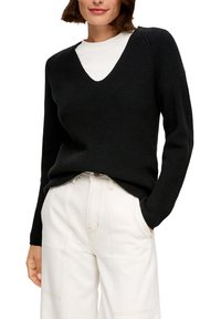 Schwarzer Strickpullover mit V-Ausschnitt und Rippstruktur, mit langen Ärmeln und lockerer Passform, kombiniert mit hellen, weiten Hosen.