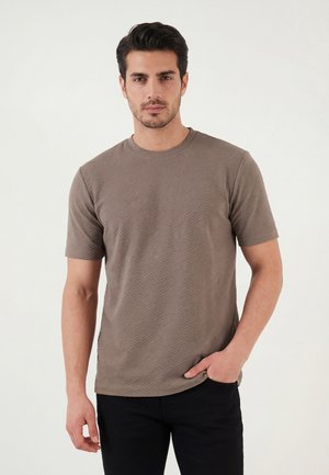Man with short dark hair wearing a textured brown short-sleeve t-shirt and black pants, standing with one hand in pocket.