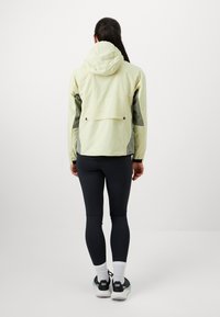 Chaqueta ligera de color verde claro con capucha, paneles laterales negros texturizados y bolsillo en la parte trasera. Combinada con mallas negras y zapatillas deportivas.