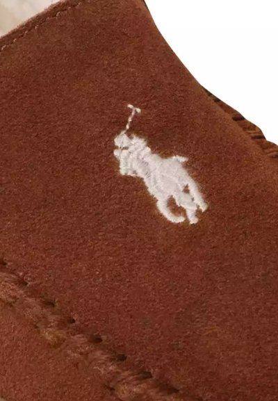 Hnědá semišová nazouvací bota s bílým vyšívaným logem. Má zaoblenou špičku a viditelné švy podél okraje. Měkká textura.