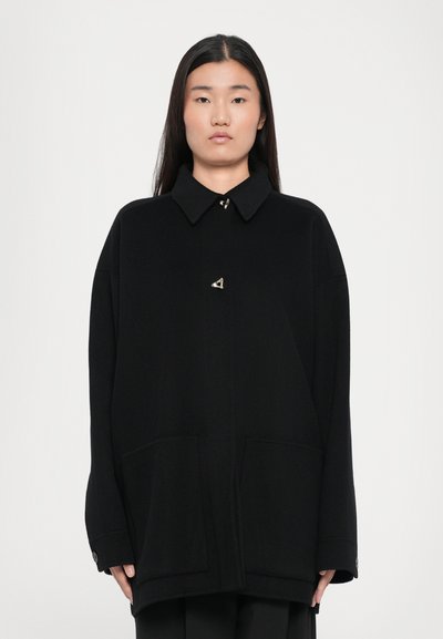 Chaqueta negra de gran tamaño con cuello con solapa, grandes bolsillos delanteros y un adorno metálico triangular. Textura suave, diseño sin costuras, estilo minimalista.