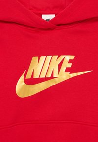 Červená mikina s kapucňou, s veľkým, lesklým zlatým logom Nike na prednej strane. Mäkká látková textúra, štandardná klokanková vrecko na spodnej strane.