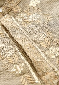 Beige tekstil med indviklet gyldent blomsterbroderi og et tekstureret mønster. Vandret dekorativ kant fremhæver designet.