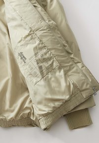 Giacca a vento beige con texture lucida e liscia, dotata di chiusura con zip e polsini a costine. Etichetta di cura visibile sulla fodera interna.