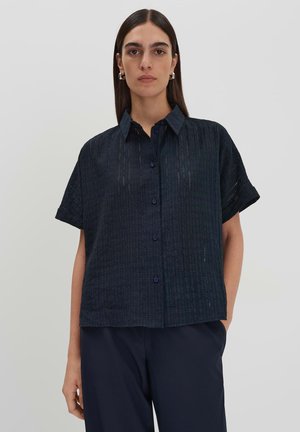 Camicia a maniche corte di colore blu scuro con un motivo texturizzato, pattina con bottoni e colletto a punta. Il materiale sembra leggero e traspirante.