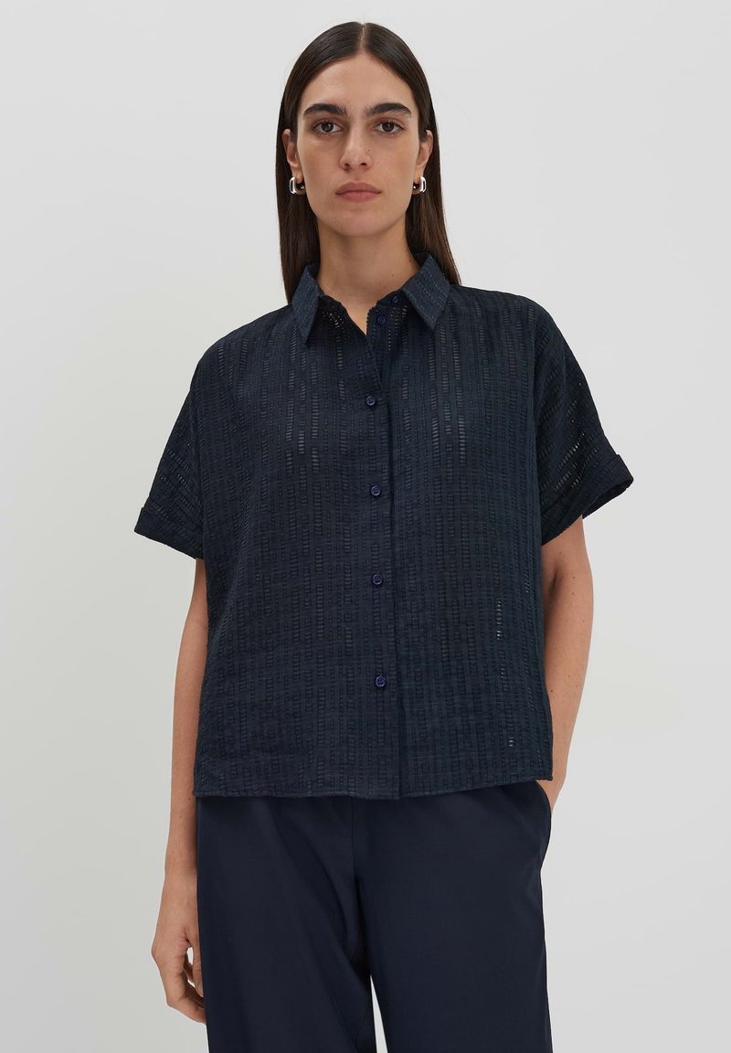 Camicia a maniche corte di colore blu scuro con un motivo texturizzato, pattina con bottoni e colletto a punta. Il materiale sembra leggero e traspirante.