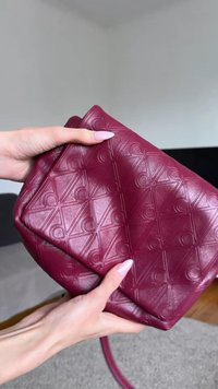 Hände halten eine gefaltete bordeauxrote Ledertasche mit einem geometrischen Muster in einem hellen Raum.