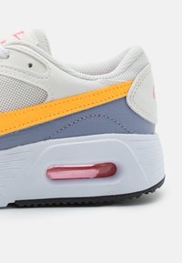 Vit sneaker med nät- och lädermaterial, med en gul accent, grå sektioner, en rosa luftenhet och strukturerad gummisula.