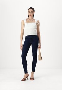 White smocked top with shoulder ties, paired with dark blue skinny jeans, brown heeled sandals, and a beige handbag. Simple design.